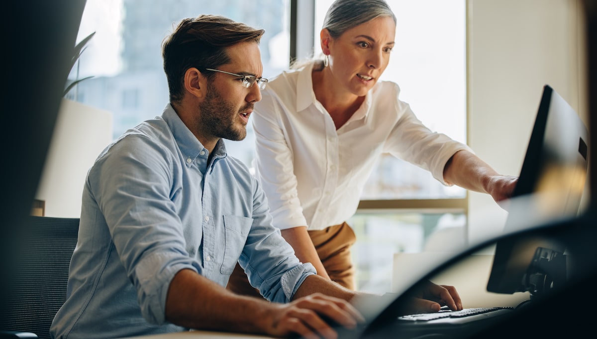 A cloud engineer shows some AWS security protocols to her colleague at his office desk.