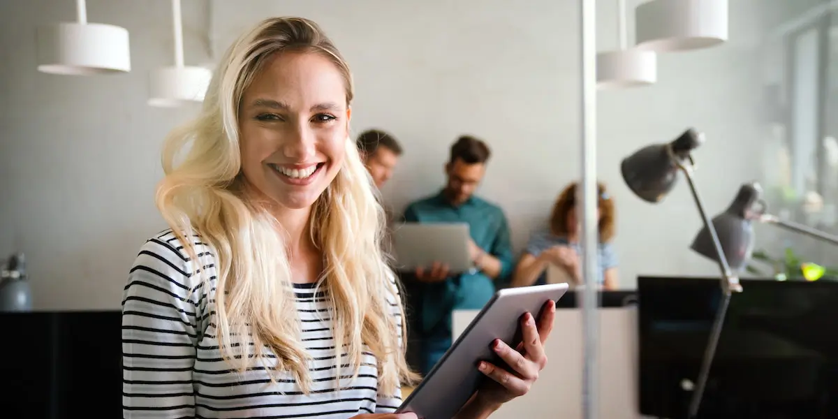 A product designer at work stands smiling at the camera while holding a tablet