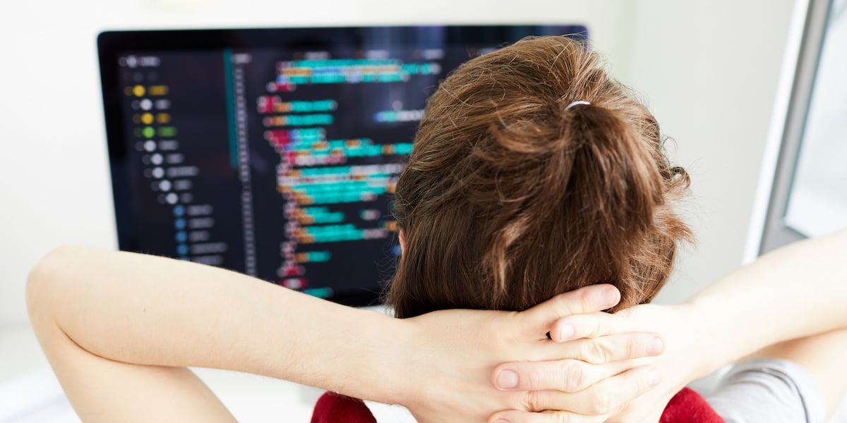 A web developer sitting at a desk with their hands behind their head, looking at a computer screen