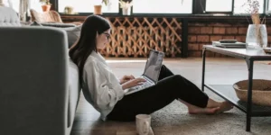 An aspiring designer sitting on the floor in their living room with a laptop and cup of coffee