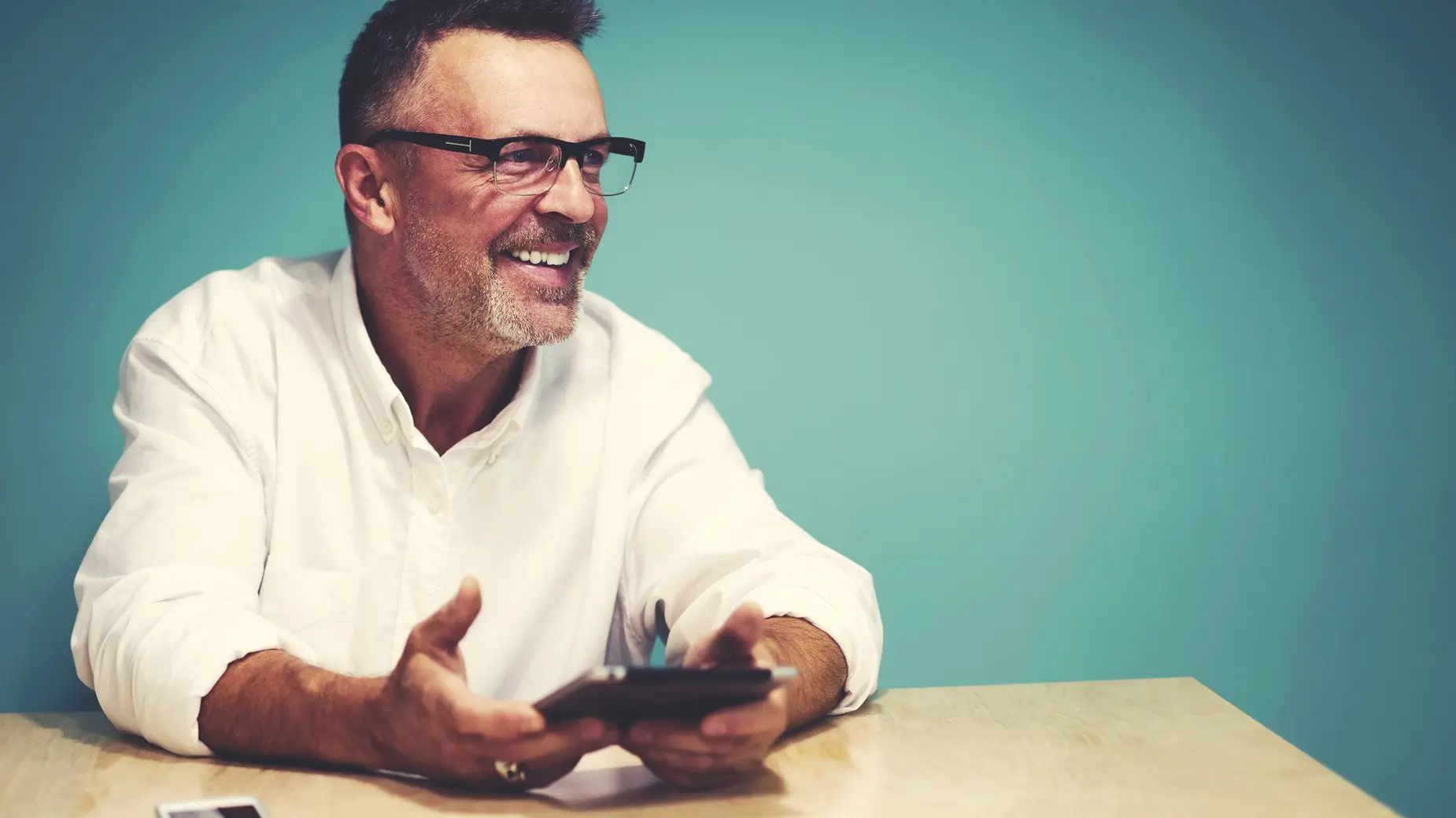 A man sitting at a table, holding a phone, in front of a teal background