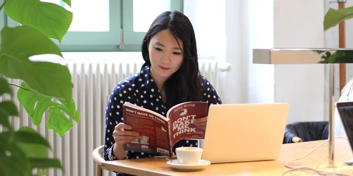 An aspiring designer ditting at a table with a laptop and cup of coffee, reading Krug's book (titled Don't Make Me Think)