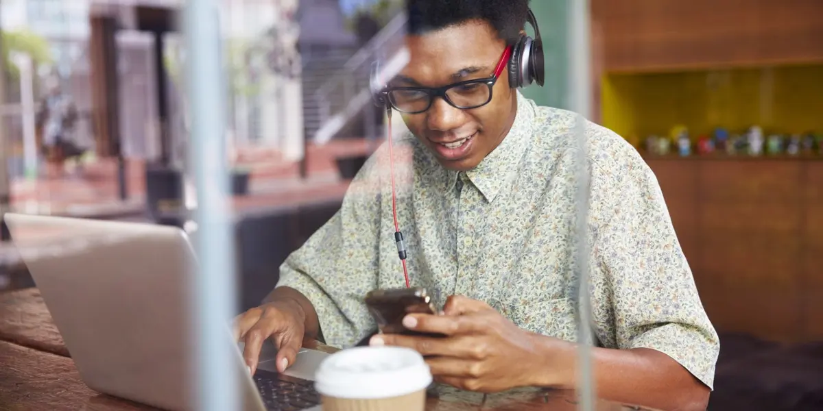 A mobile web developer wearing headphones, sitting in front of his laptop and scrolling through a smartphone