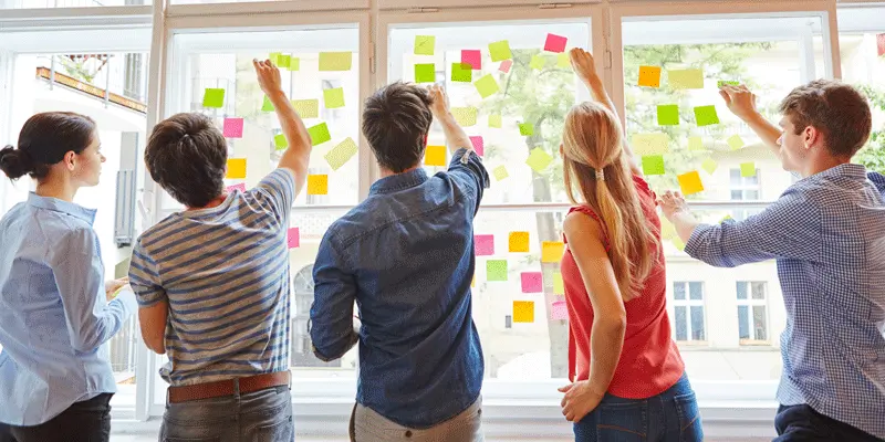 Five designers stand at a glass wall using sticky notes for ideation