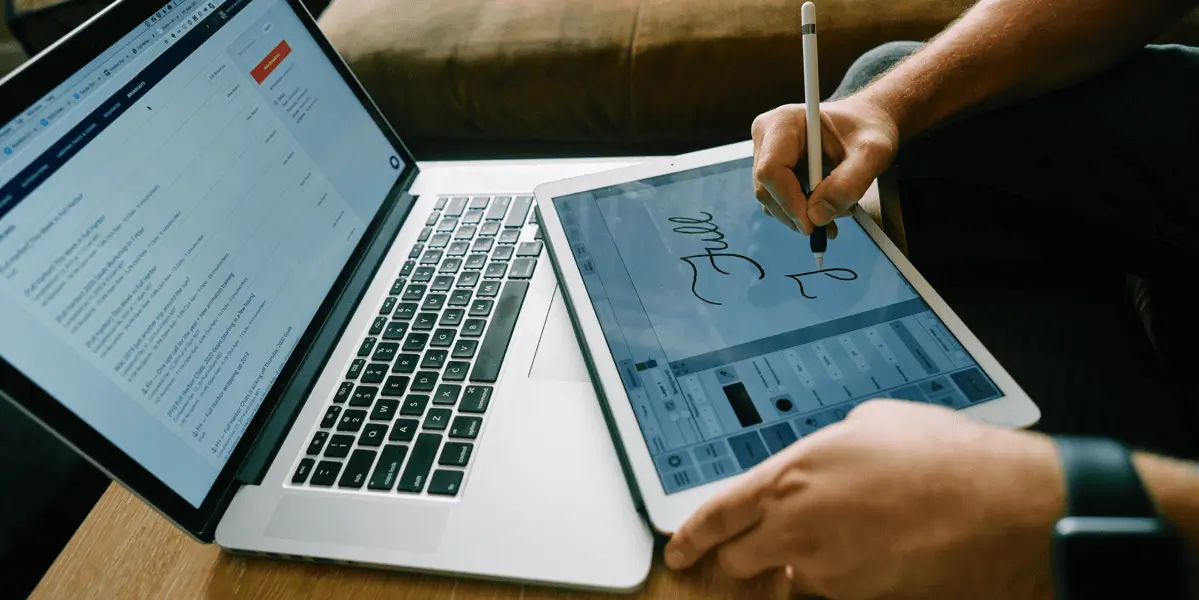 A designer sketching on a tablet with a laptop in front of them