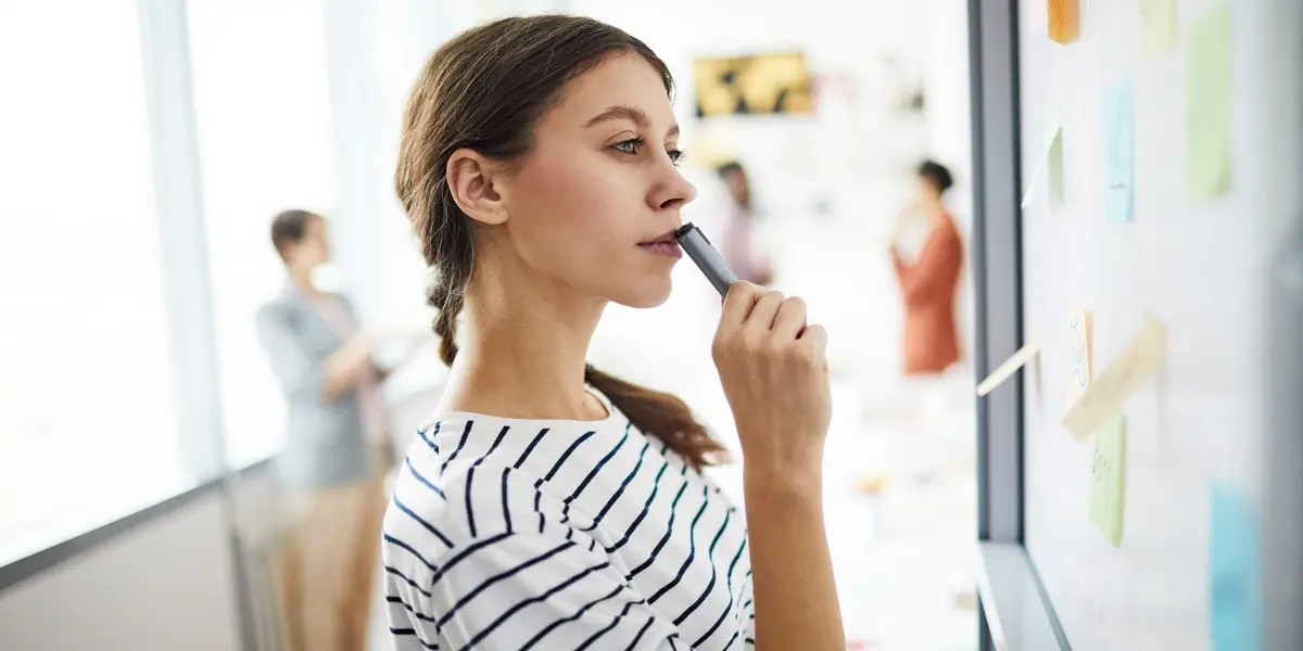 An aspiring UX designer at a whiteboard, pressing a closed marker to her lips, deep in thought