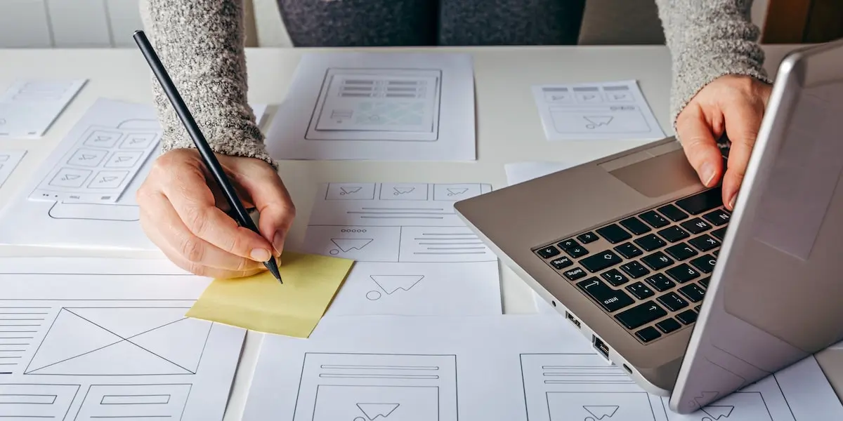 A UX designer working at a table with a laptop, writing ideas on a sticky note