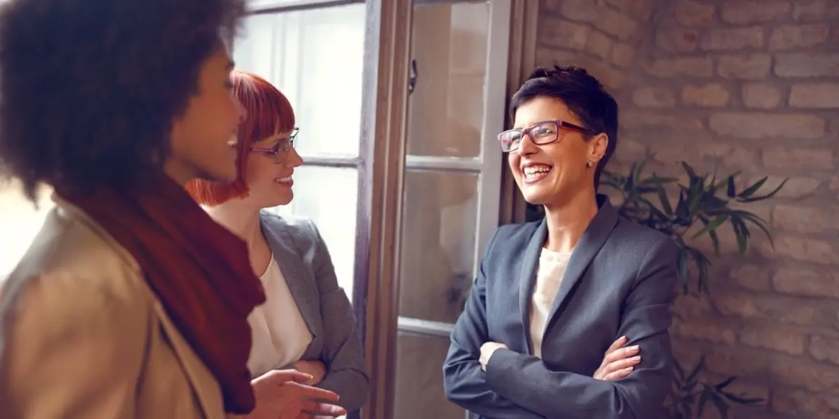 Three women in business attire, standing near a window with a brick wall and plants in the background