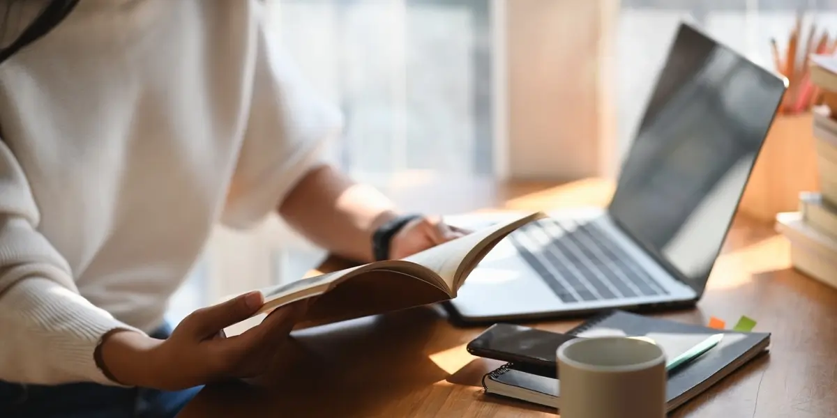 a UX designer sitting at a table with an open laptop, flipping through a notebook