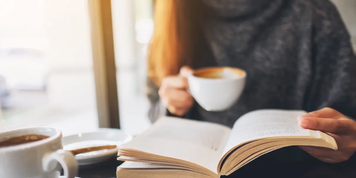 A UX writer sitting at a table near a window, drinking a coffee and reading