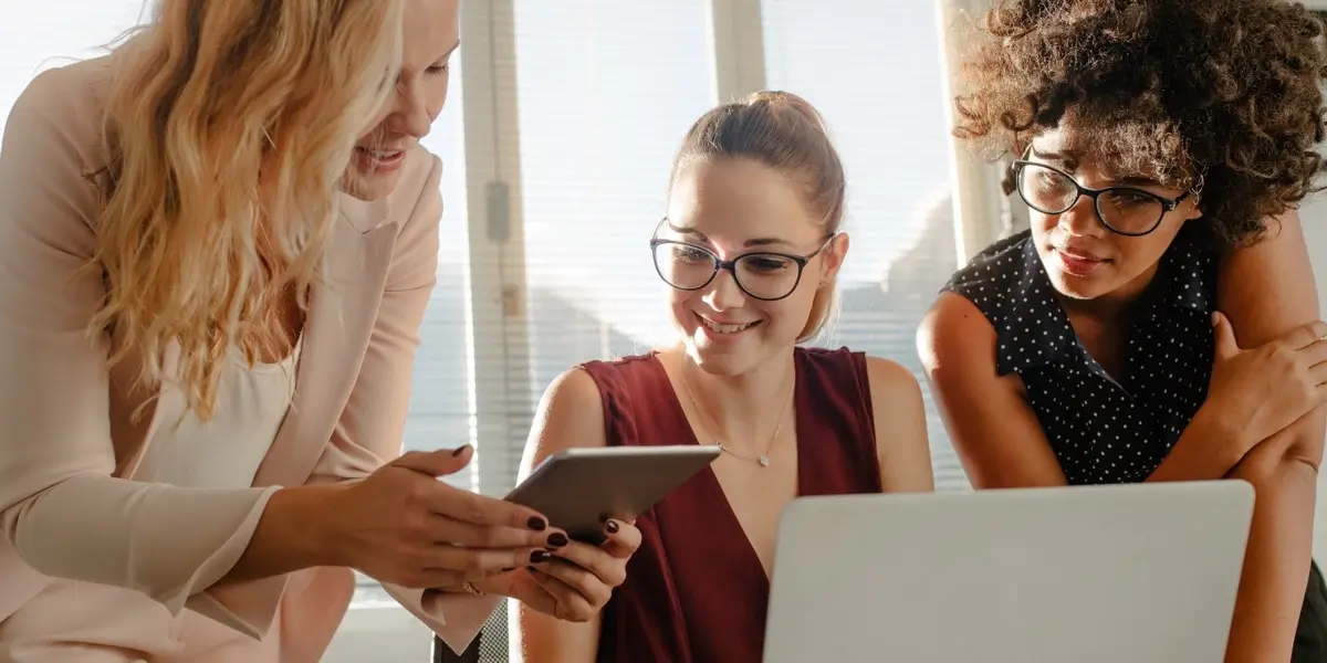 Three women gathered around a laptop and tablet, looking at projects notes