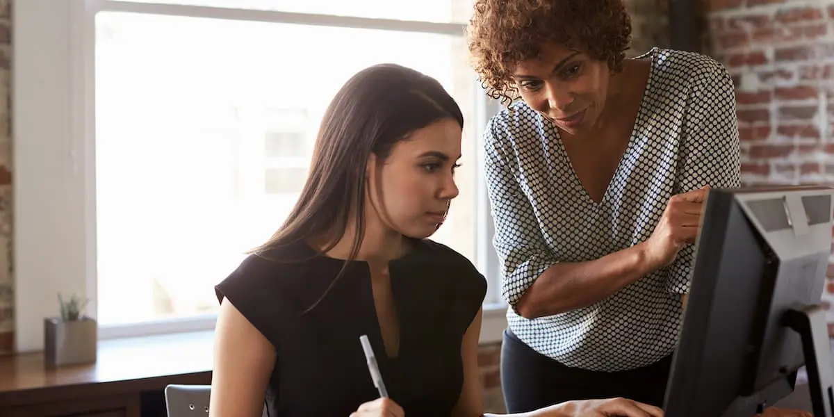 An aspiring UX designer sitting at a desk, as their UX mentor looks over a project and offers feedback.