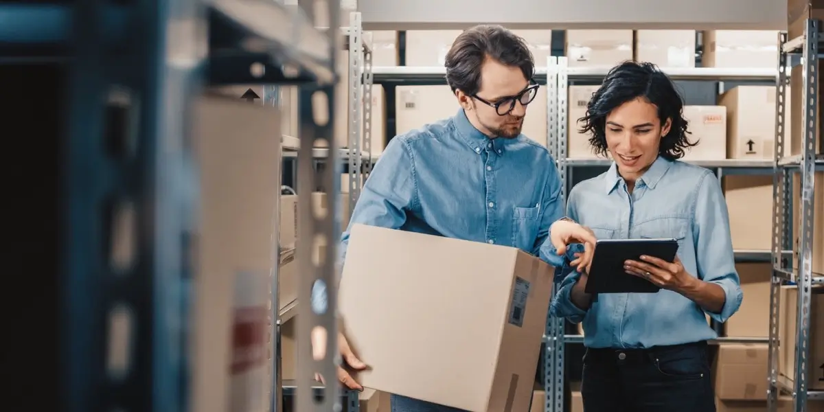 Two people standing in a warehouse, one looking at a tablet, the other holding a box for shipment