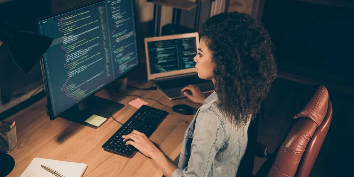 A web developer sits at a desk in her home office with multiple screens of code in front of her.