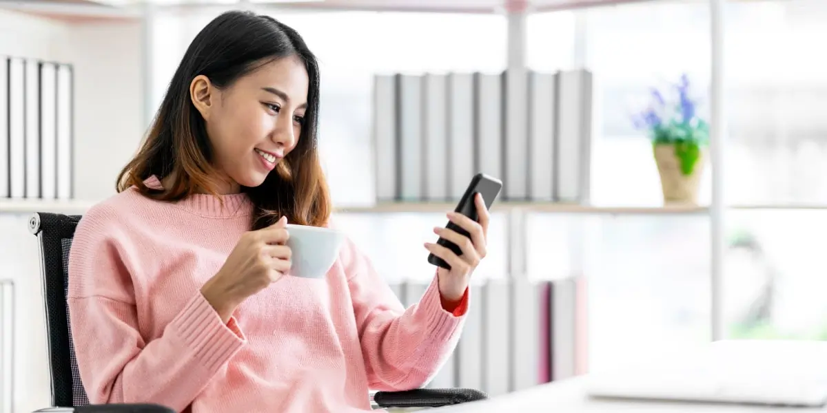 An aspiring marketer sitting at a desk, browsing social media jobs on a phone, drinking a cup of coffee