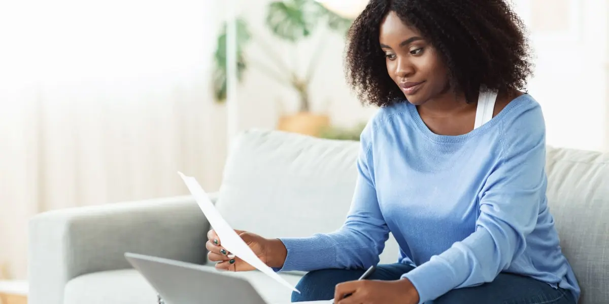 A digital marketing degree student sitting on a sofa, looking at a laptop and holding a sheet of paper