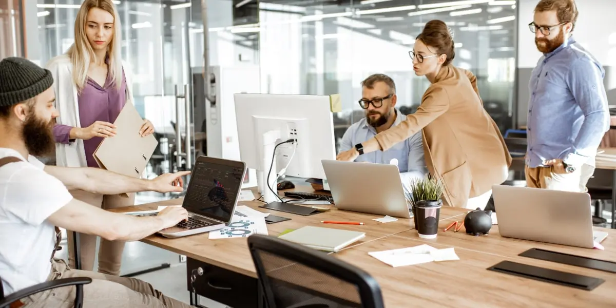 A team of web developers sit at a table of laptops and at a desktop pc in a startup planning how to use the django framework for a project