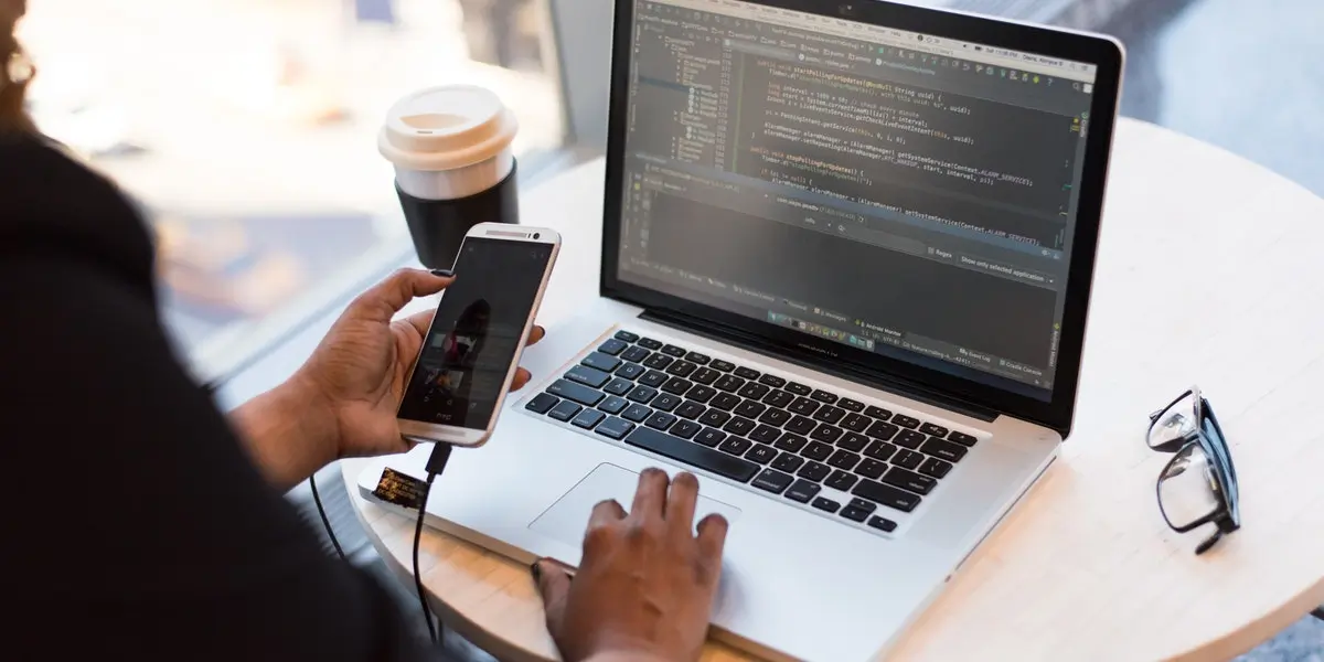 A programmer works on code on their laptop and checks their smartphone doing mobile app development in a café.