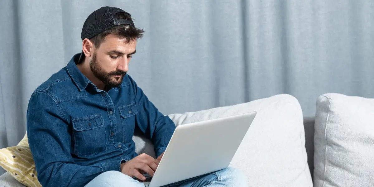 A marketing student sitting on a sofa, wearing a cap, researching the average digital marketing specialist salary on a laptop