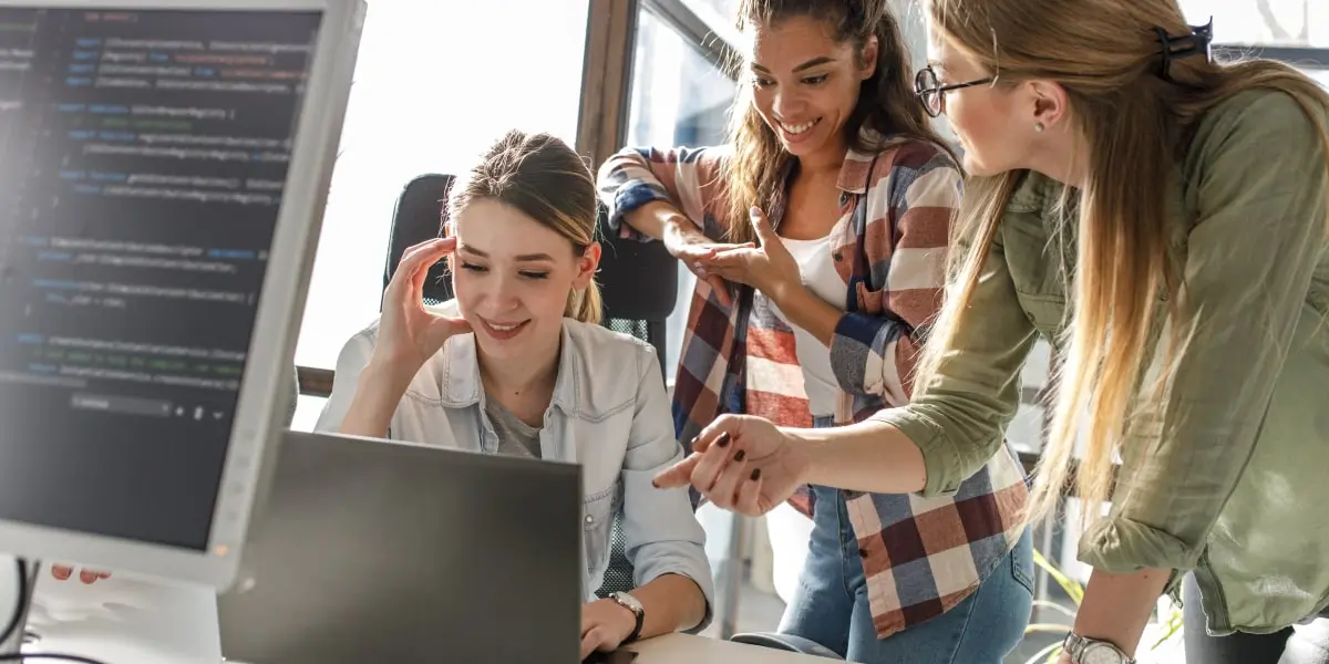 A senior software engineer helps two junior developers in an office with some code on a laptop.