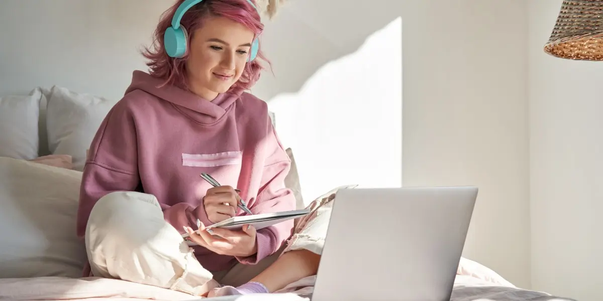 A woman sits on her bed with her laptop studying at an online coding school.