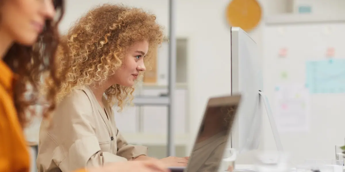 A full-stack developer sitting working at a computer in a startup office with her colleague in the foreground.