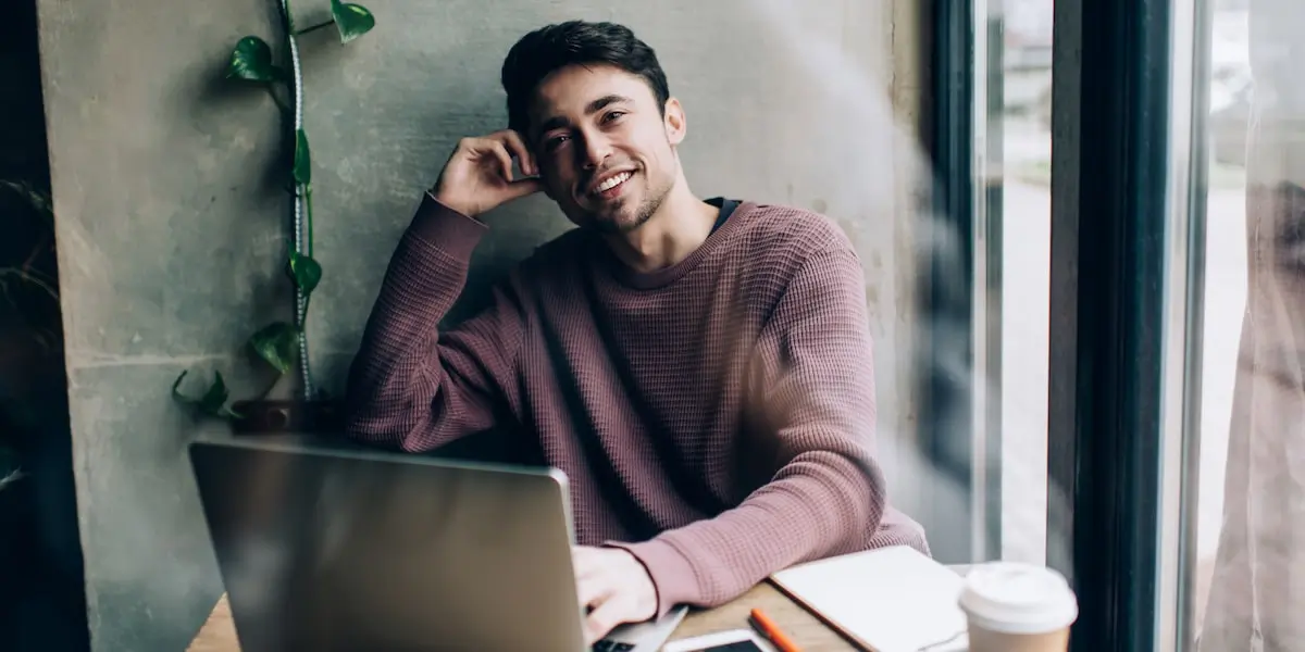 A man sits in a cafe with a laptop and notepad wondering is coding hard to learn.