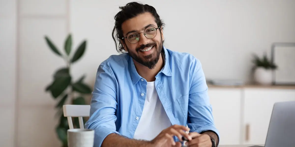 A backend developer with braces sits at a table with a laptop, smiling at the camera.