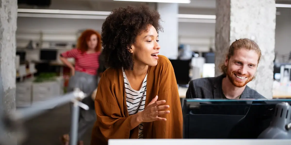 A woman discusses a project for her product manager portfolio with her colleague at a computer.