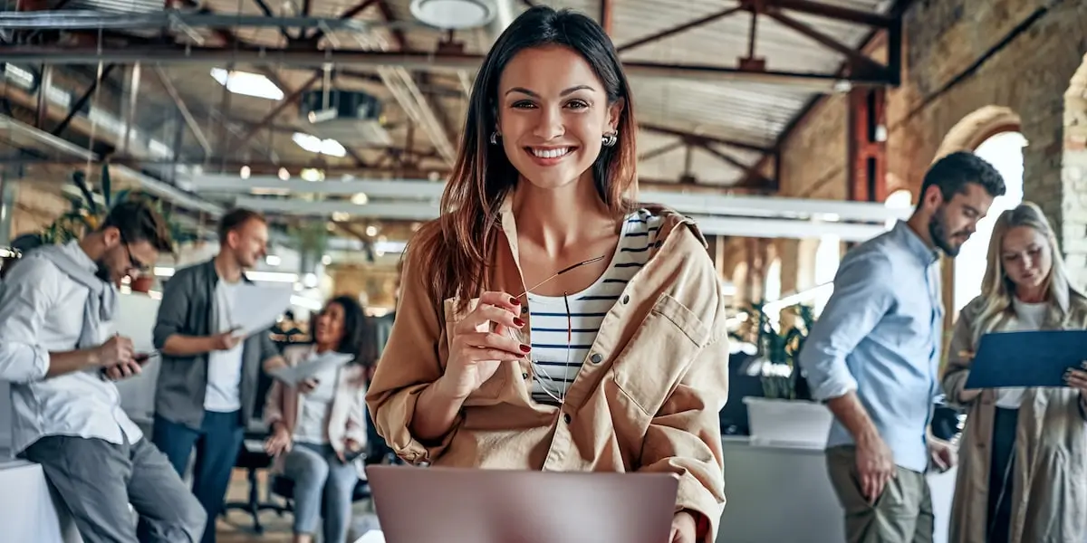 A scrum master looks at the camera holding a laptop at a scrum meeting in an office.