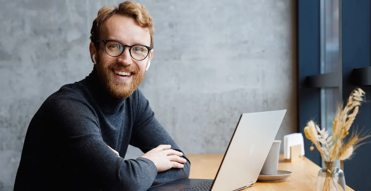 Software engineer in a cafe smiles at the camera after learning what is the MERN stack on his laptop.