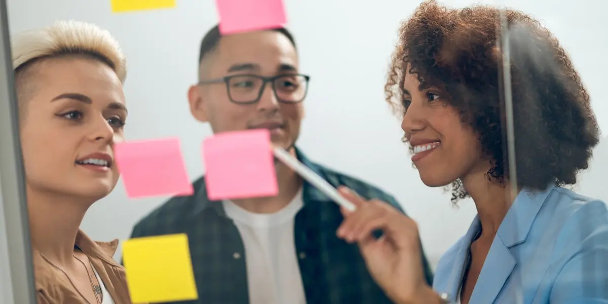 Scrum master shows two other employees what is a scrum meeting using sticky notes on a board.