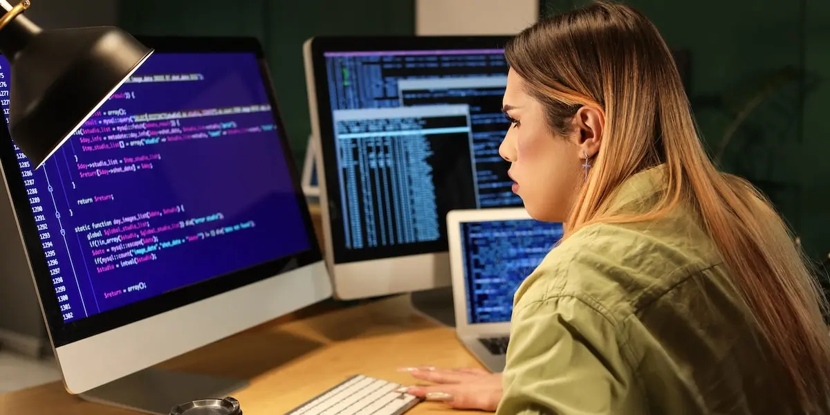 A programmer sits at two desktop screens and a laptop screen comparing angular vs react code on them.