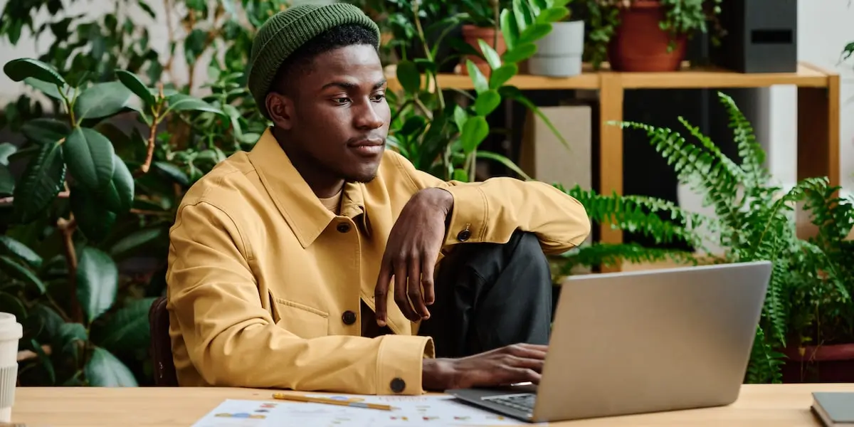 A man sits in a leafy coffee shop on a laptop learning what is coding.