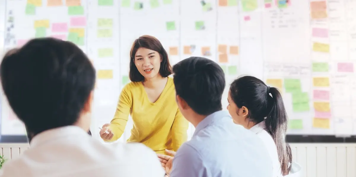 A scrum master explains what is a scrum team to her new colleagues with a Kanban board in the background.