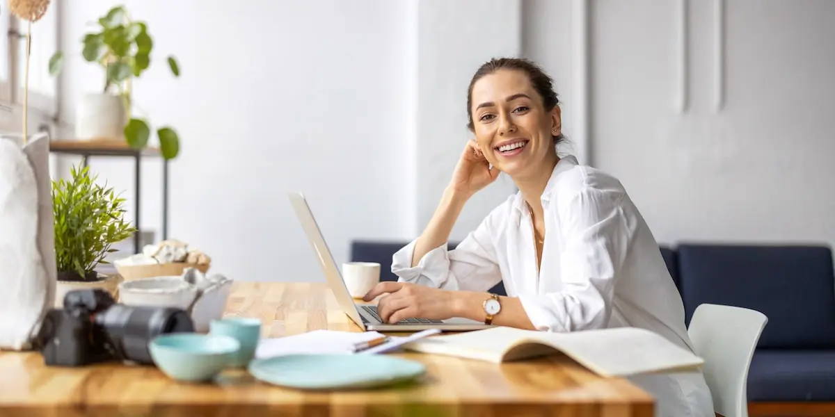 A woman learning coding for beginners sits at her laptop in a bright room smiling at the camera.