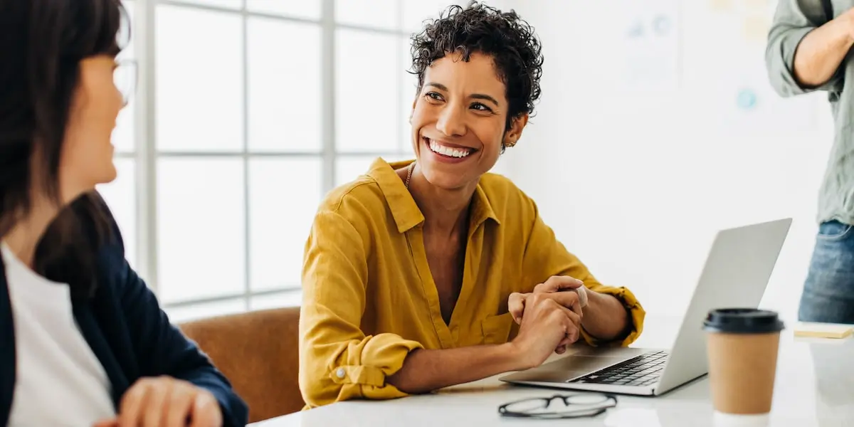 A product analyst sits in an office meeting with her laptop smiling.