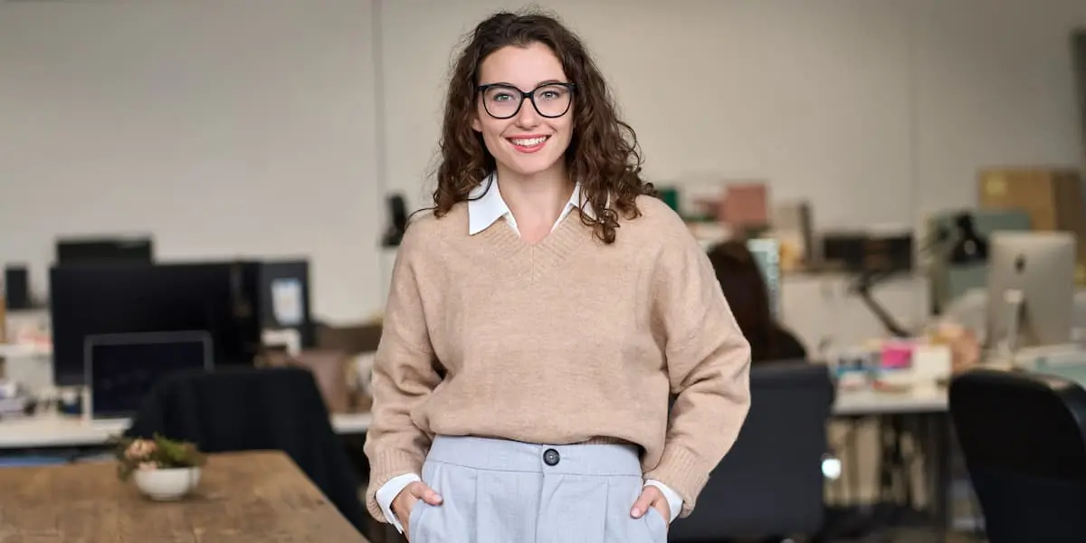 a young brunette stands smiling at the camera with their back to the office and hands in their pockets