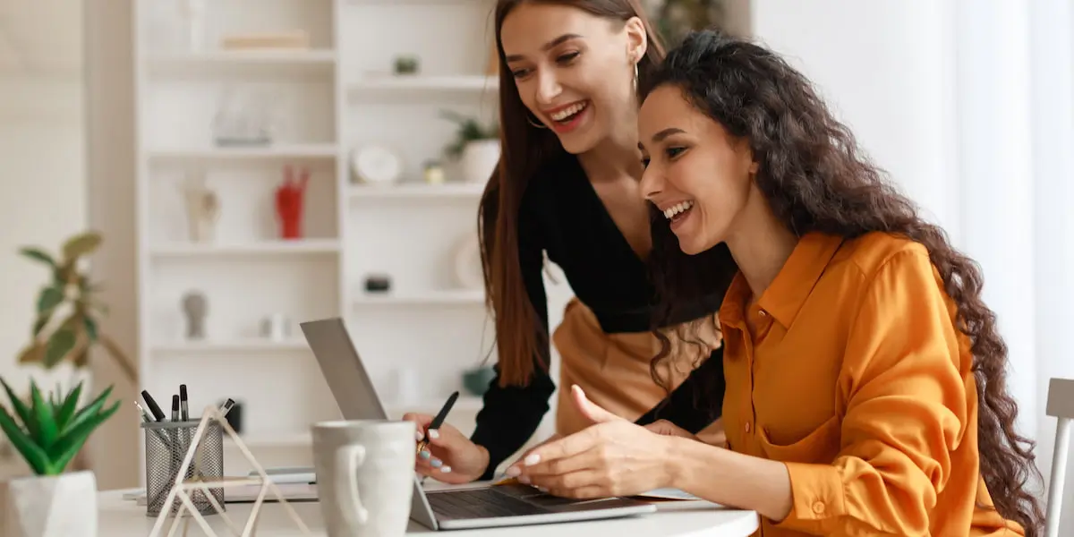 Two young ladies, one standing and one sitting, smiling at a laptop.