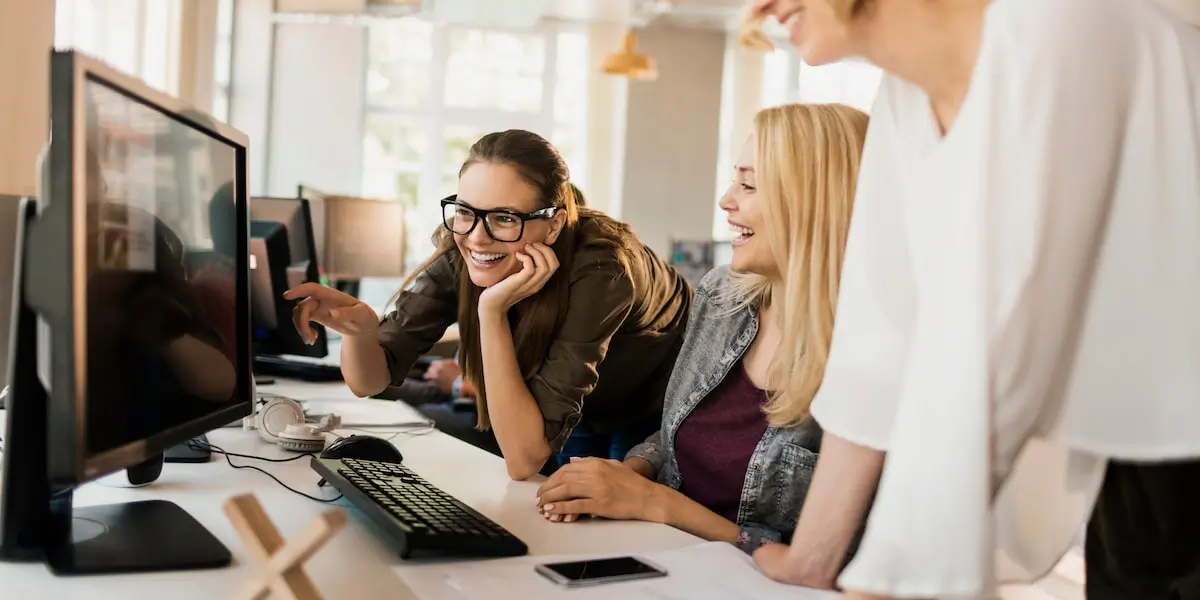 A community manager with her two teamates points at a desktop computer screen while all three laugh