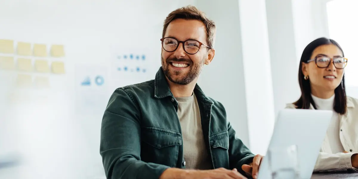 Two digital marketing experts sit at laptops with a focus on the smiling bearded male wearing glasses.