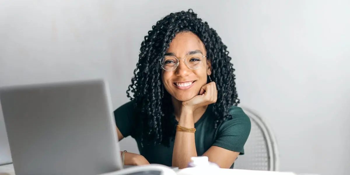 A young woman smiles at the computer with her head resting on her hand