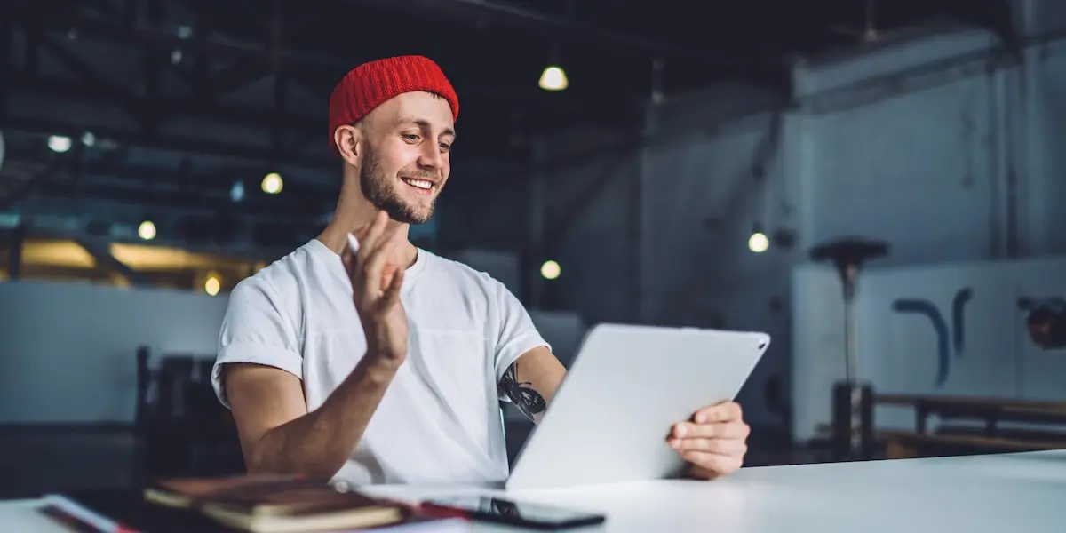 Young male designer in red beanie waving at his tablet