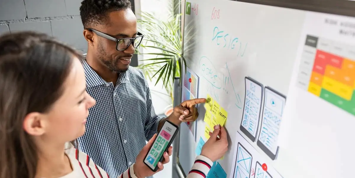 Two UX designers work on writing their UX user stories at a whiteboard showing wireframes and prototypes.